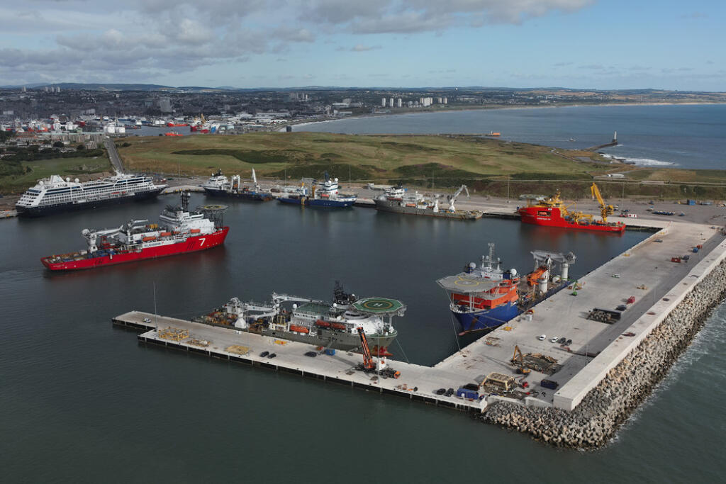 Aerial view of Port of Aberdeen&#039;s bustling South Harbour with red and blue vessels docked along its various quaysides. A large cruise ship is seen on South Harbour&#039;s Balmoral Quay, with the port&#039;s North Harbour, the Girdleness Lighthouse and Aberdeen City Centre in the distance under a partly cloudy sky.